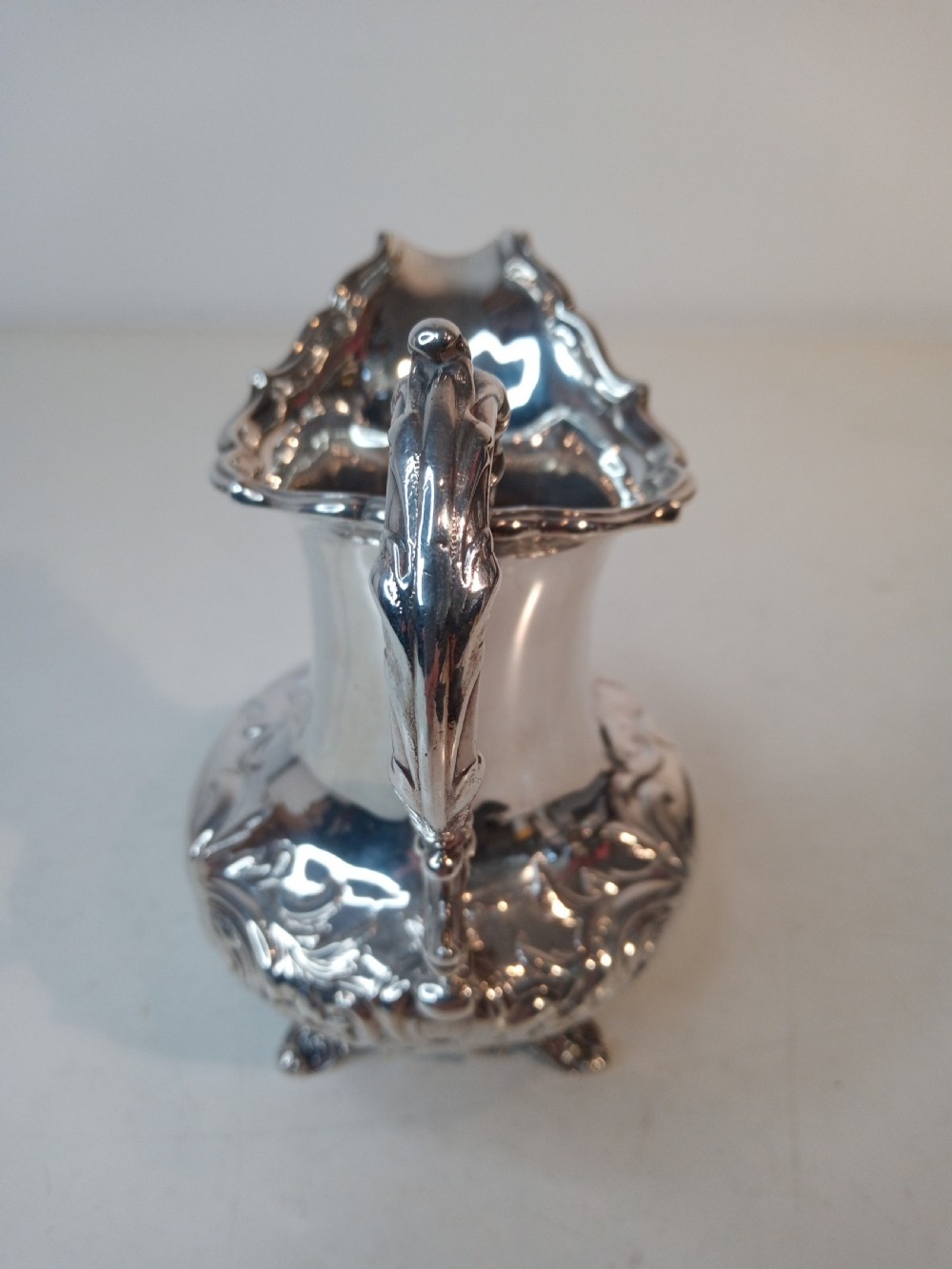 A Victorian hallmarked sterling silver jug with intricate scroll chasing, a shaped pour spout, a decorative handle, and scroll feet. View IV