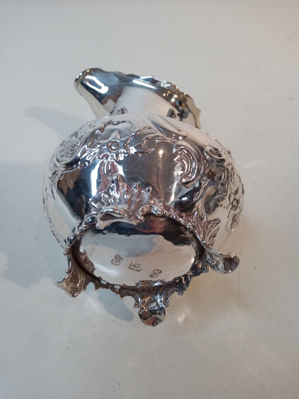 A Victorian hallmarked sterling silver jug with intricate scroll chasing, a shaped pour spout, a decorative handle, and scroll feet. View VI