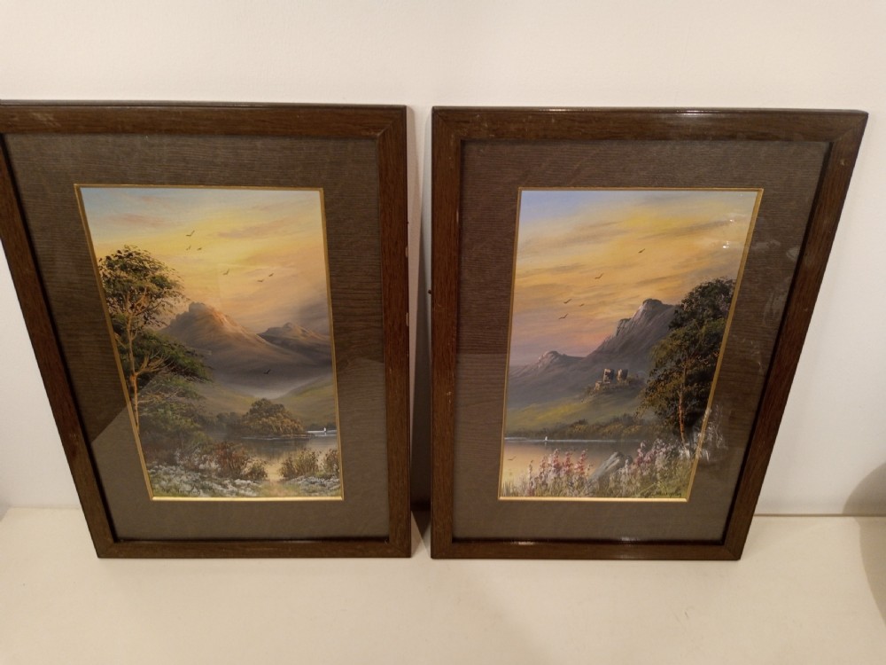 A pair of framed early 20th-century gouache landscape paintings depicting mountainous scenes with bodies of water framed in dark wooden mounts and frames.