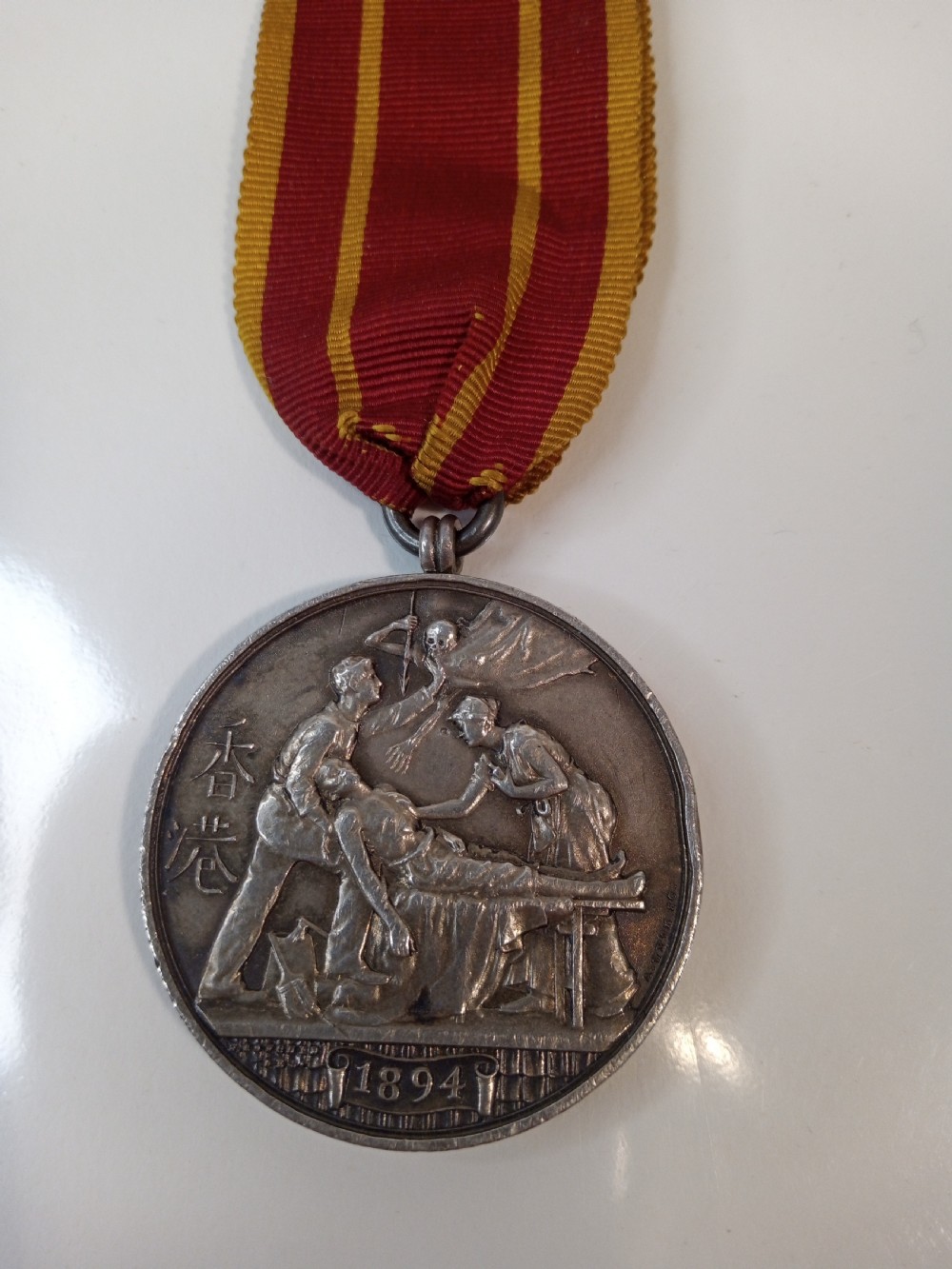 An antique silver circular service medal dated 1894 featuring relief figures and Chinese characters, suspended from a red and yellow striped silk ribbon.