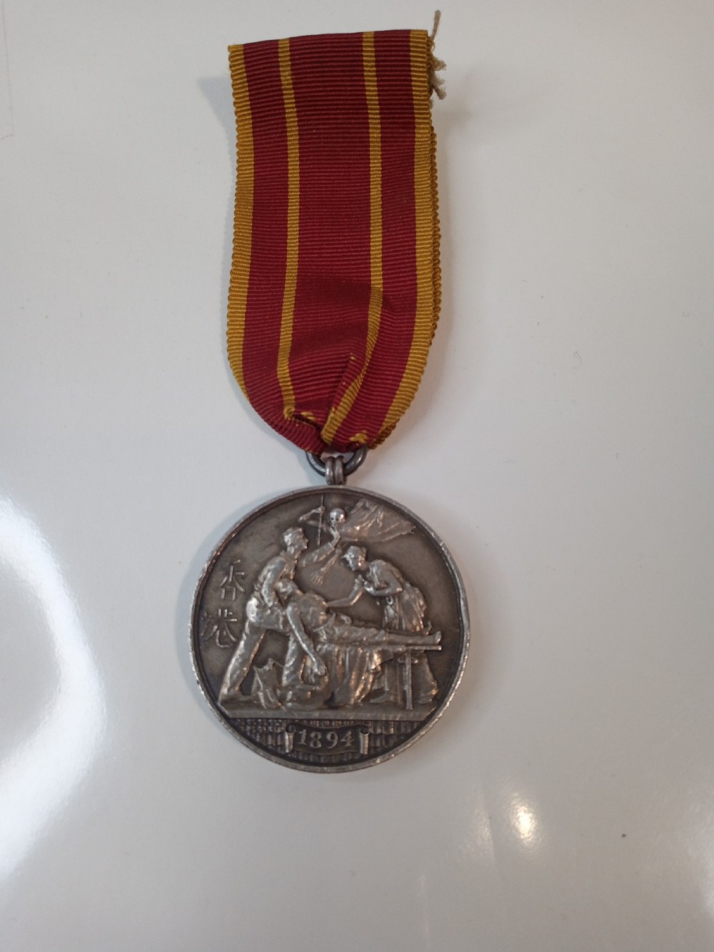 An antique silver circular service medal dated 1894 featuring relief figures and Chinese characters, suspended from a red and yellow striped silk ribbon. View II
