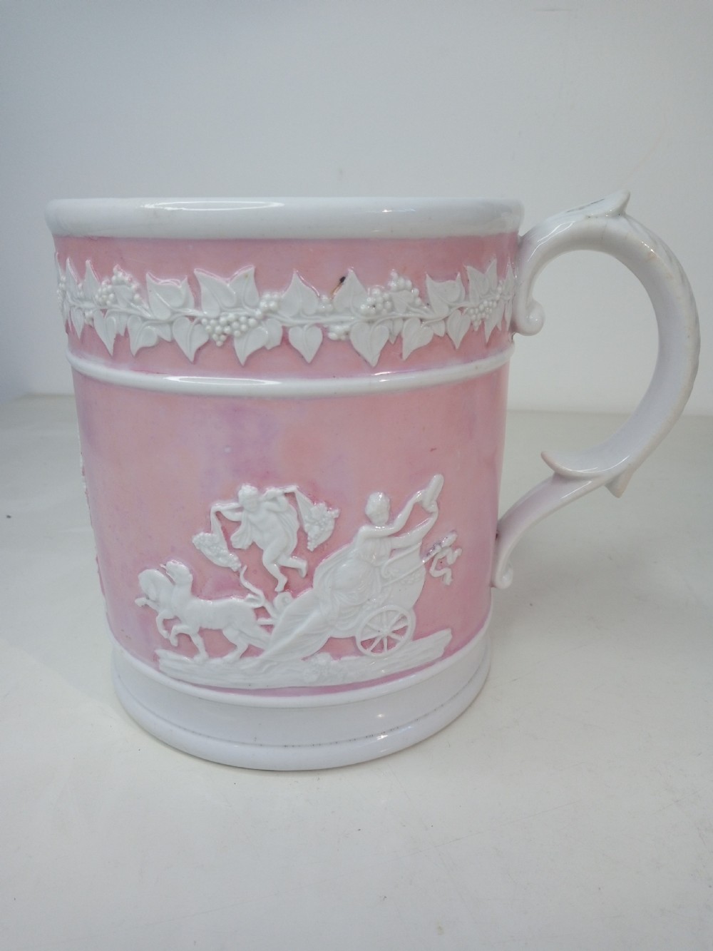 A cylindrical pink porcelain tankard featuring white moulded relief figures and a scrolled handle against a plain background.