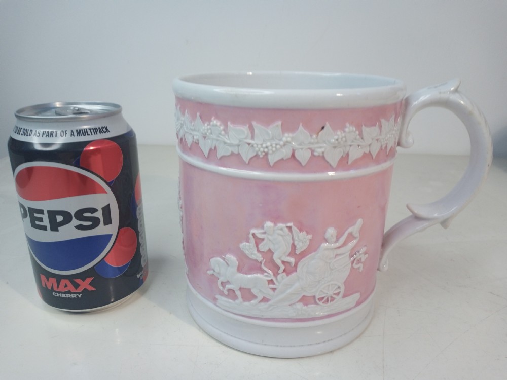 A cylindrical pink porcelain tankard featuring white moulded relief figures and a scrolled handle against a plain background. View II