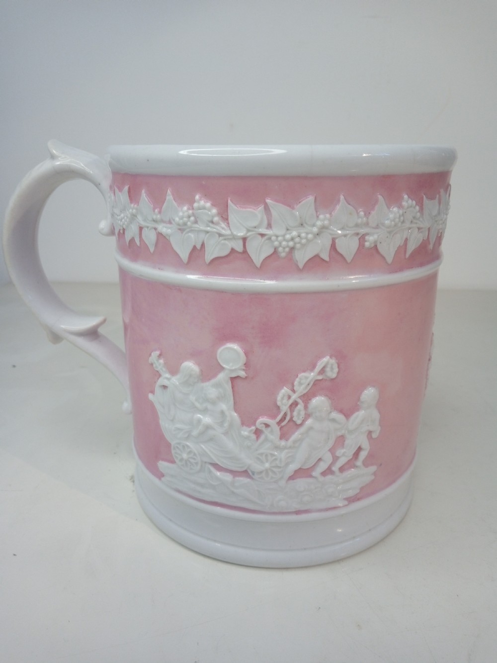 A cylindrical pink porcelain tankard featuring white moulded relief figures and a scrolled handle against a plain background. View III