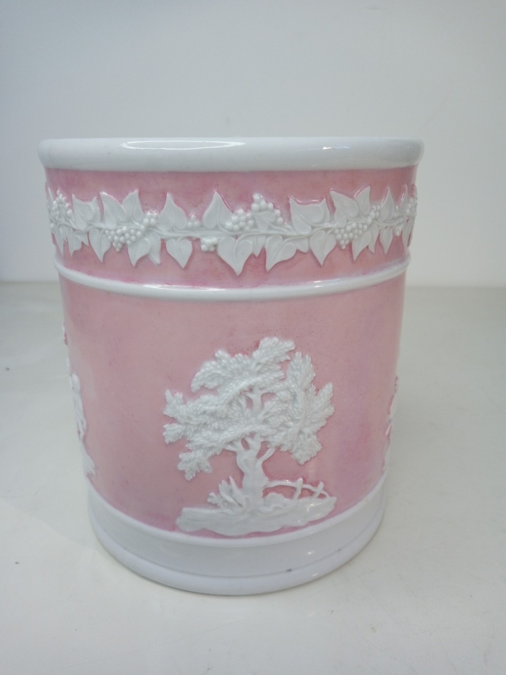 A cylindrical pink porcelain tankard featuring white moulded relief figures and a scrolled handle against a plain background. View IV