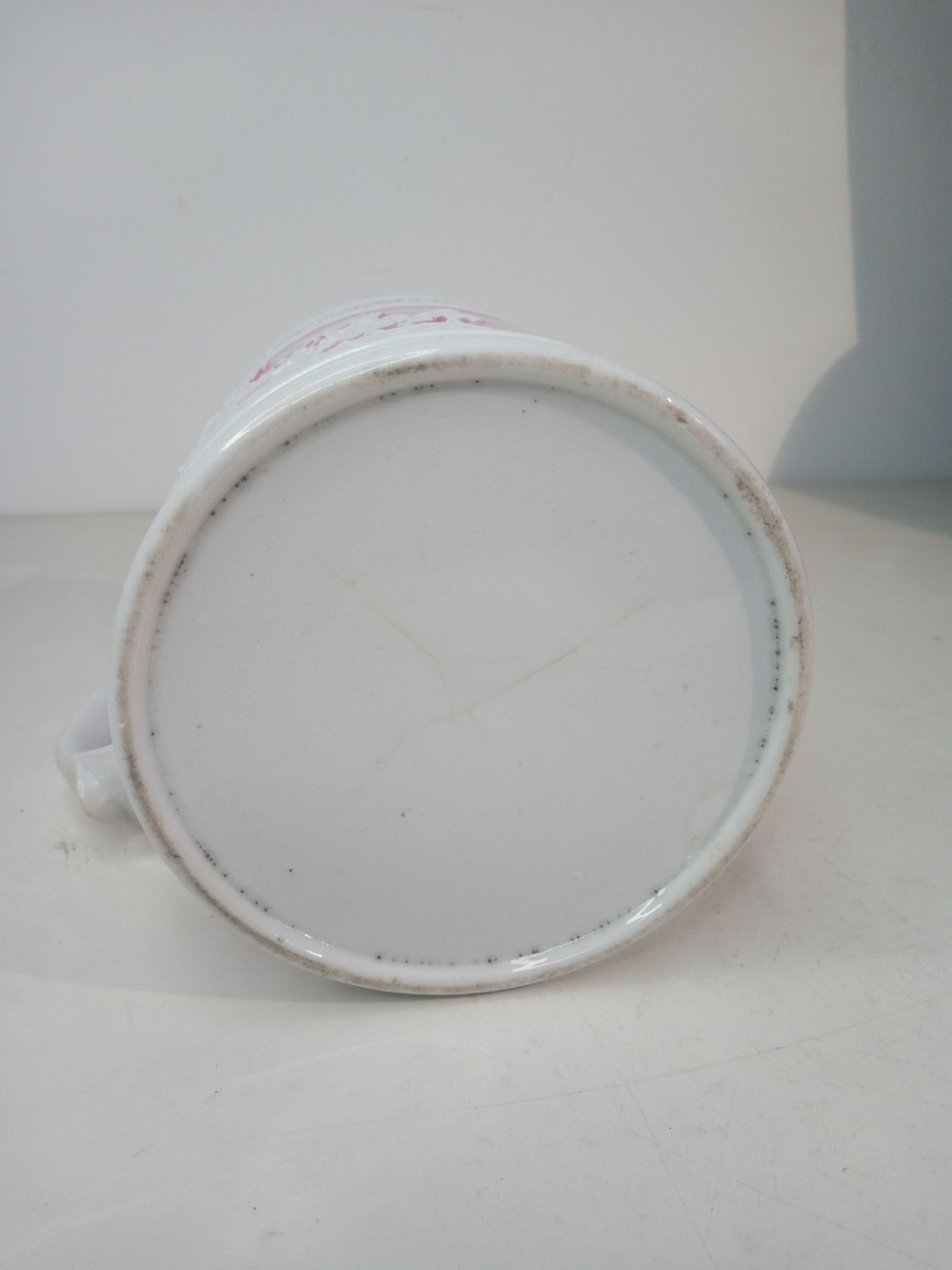 A cylindrical pink porcelain tankard featuring white moulded relief figures and a scrolled handle against a plain background. View V