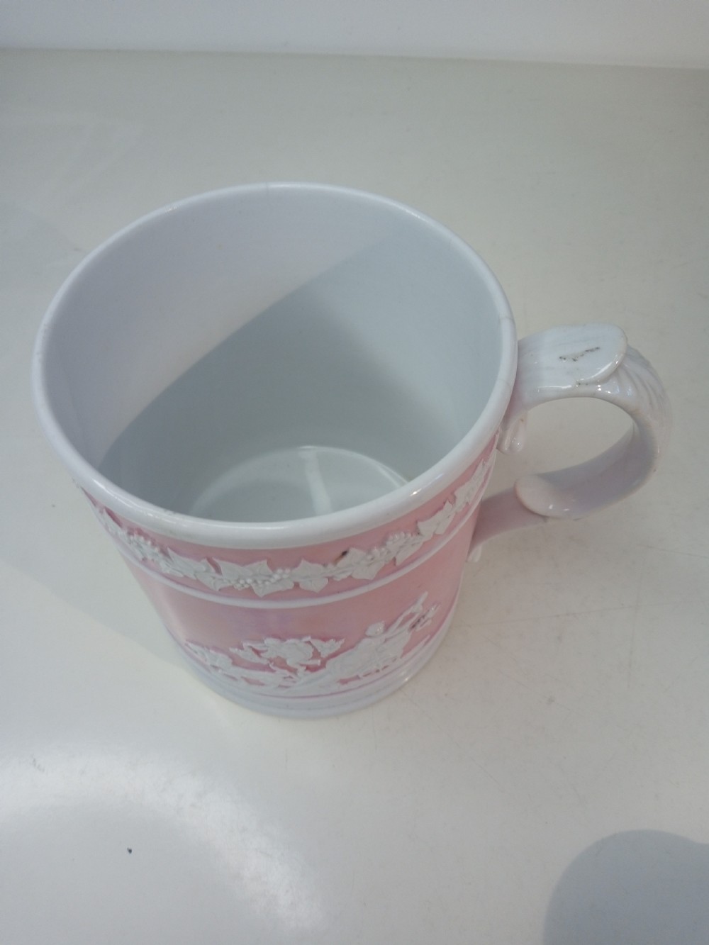 A cylindrical pink porcelain tankard featuring white moulded relief figures and a scrolled handle against a plain background. View VII
