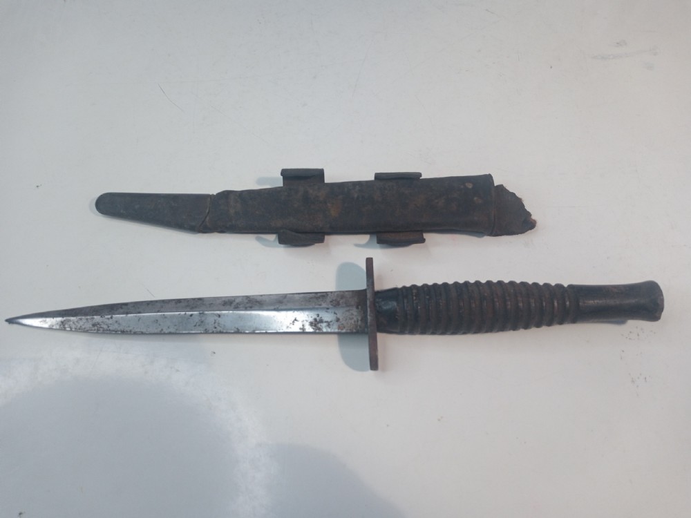A World War II double-edged commando fighting knife with a ribbed handle positioned below its dark leather scabbard.