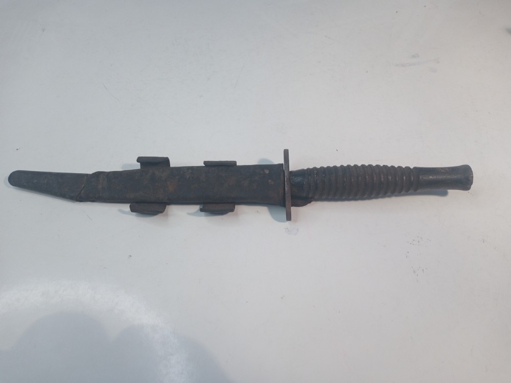 A World War II double-edged commando fighting knife with a ribbed handle positioned below its dark leather scabbard. View II