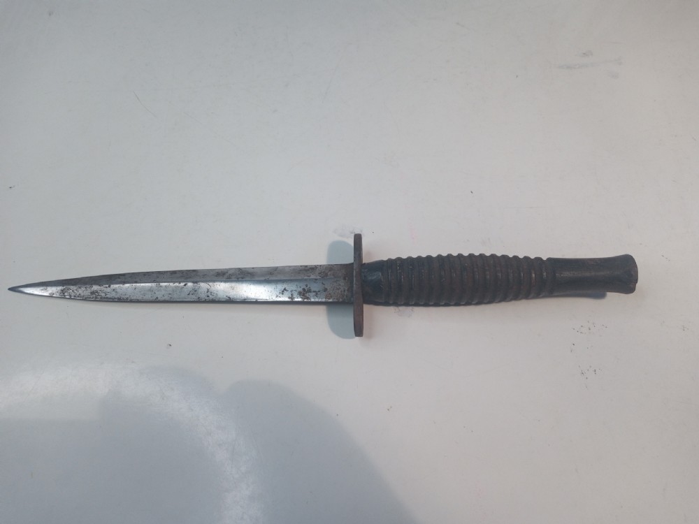 A World War II double-edged commando fighting knife with a ribbed handle positioned below its dark leather scabbard. View III