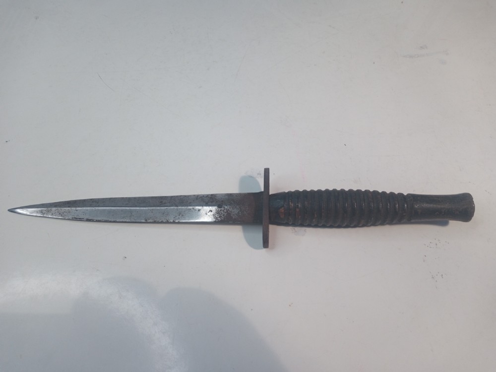 A World War II double-edged commando fighting knife with a ribbed handle positioned below its dark leather scabbard. View IV