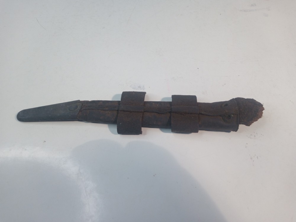 A World War II double-edged commando fighting knife with a ribbed handle positioned below its dark leather scabbard. View VI