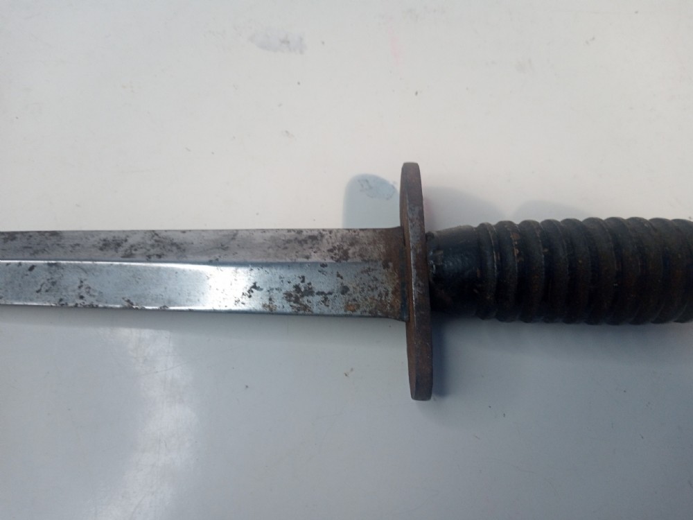 A World War II double-edged commando fighting knife with a ribbed handle positioned below its dark leather scabbard. View VII