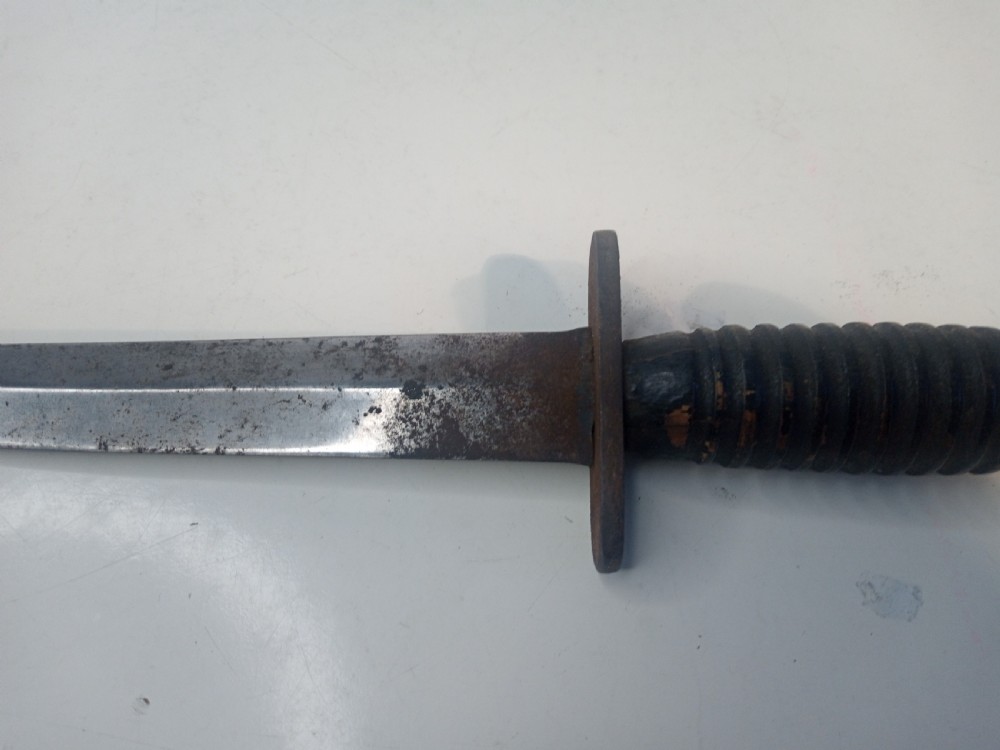 A World War II double-edged commando fighting knife with a ribbed handle positioned below its dark leather scabbard. View VIII