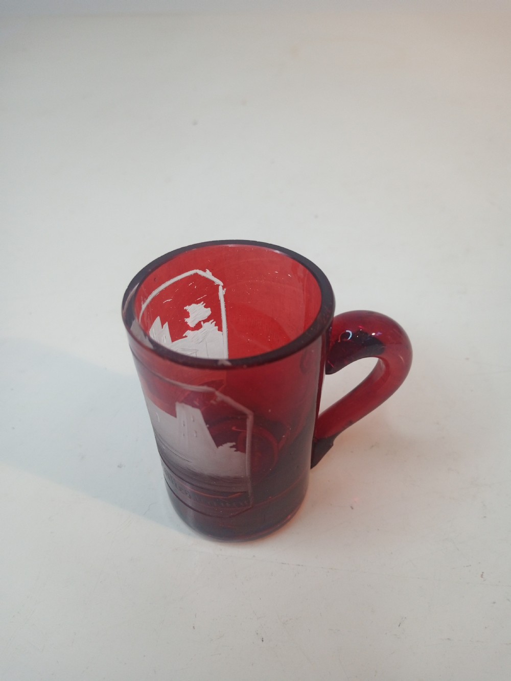 A late Victorian miniature ruby glass tankard with an applied red handle and a white etched architectural motif on the body.