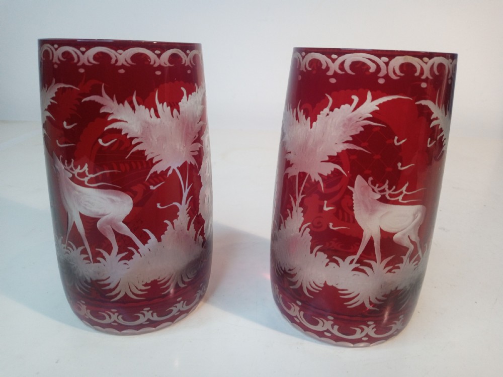 A pair of ruby red glass tumblers featuring white wheel-engraved stag and forest motifs with decorative scrollwork borders.
