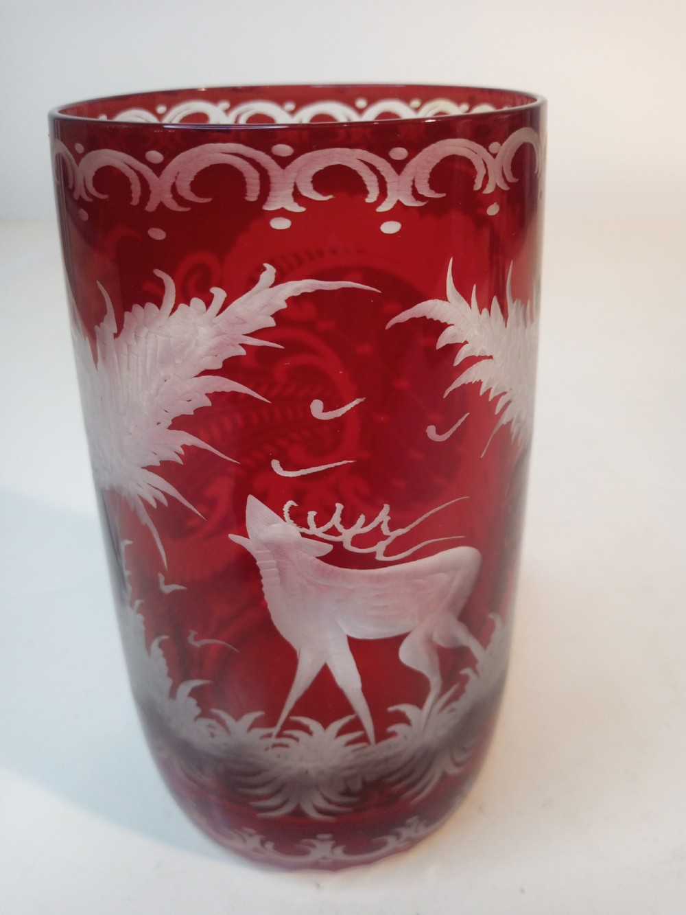 A pair of ruby red glass tumblers featuring white wheel-engraved stag and forest motifs with decorative scrollwork borders. View X