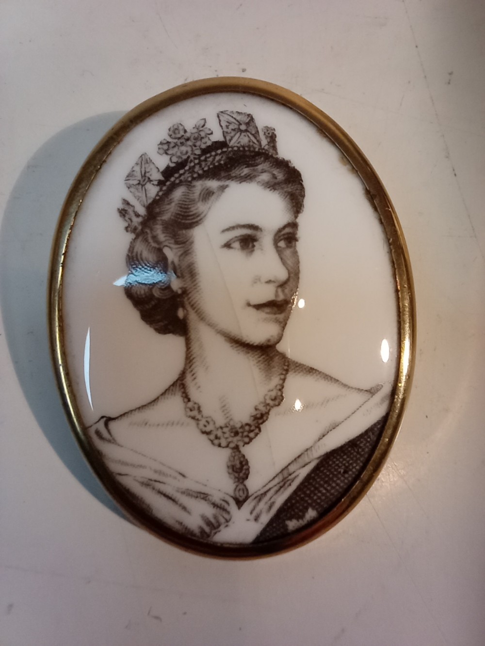 An oval Royal Worcester porcelain brooch featuring a black and white transfer-printed portrait of Queen Elizabeth II wearing a tiara, set inside a smooth gilt metal rim.