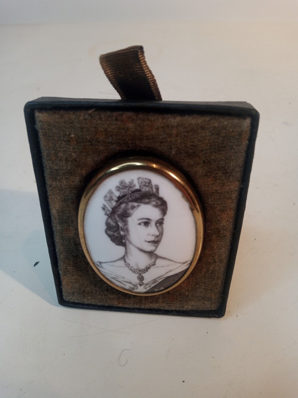 An oval Royal Worcester porcelain brooch featuring a black and white transfer-printed portrait of Queen Elizabeth II wearing a tiara, set inside a smooth gilt metal rim. View II