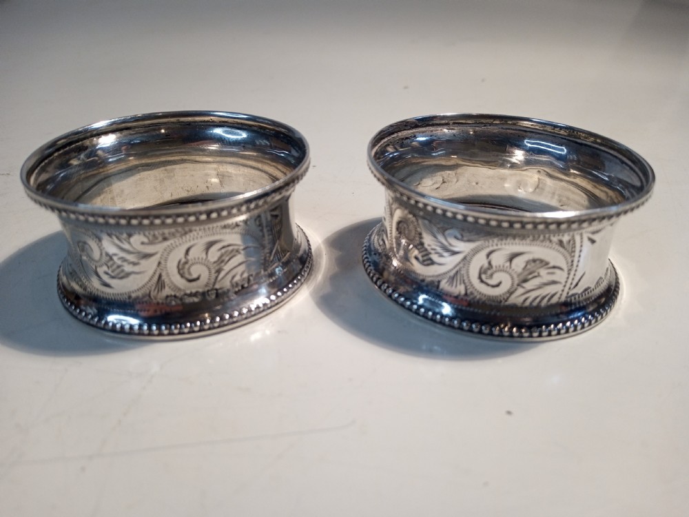 A pair of engraved sterling silver napkin rings with beaded rims and hallmarked details resting on a light surface. View II