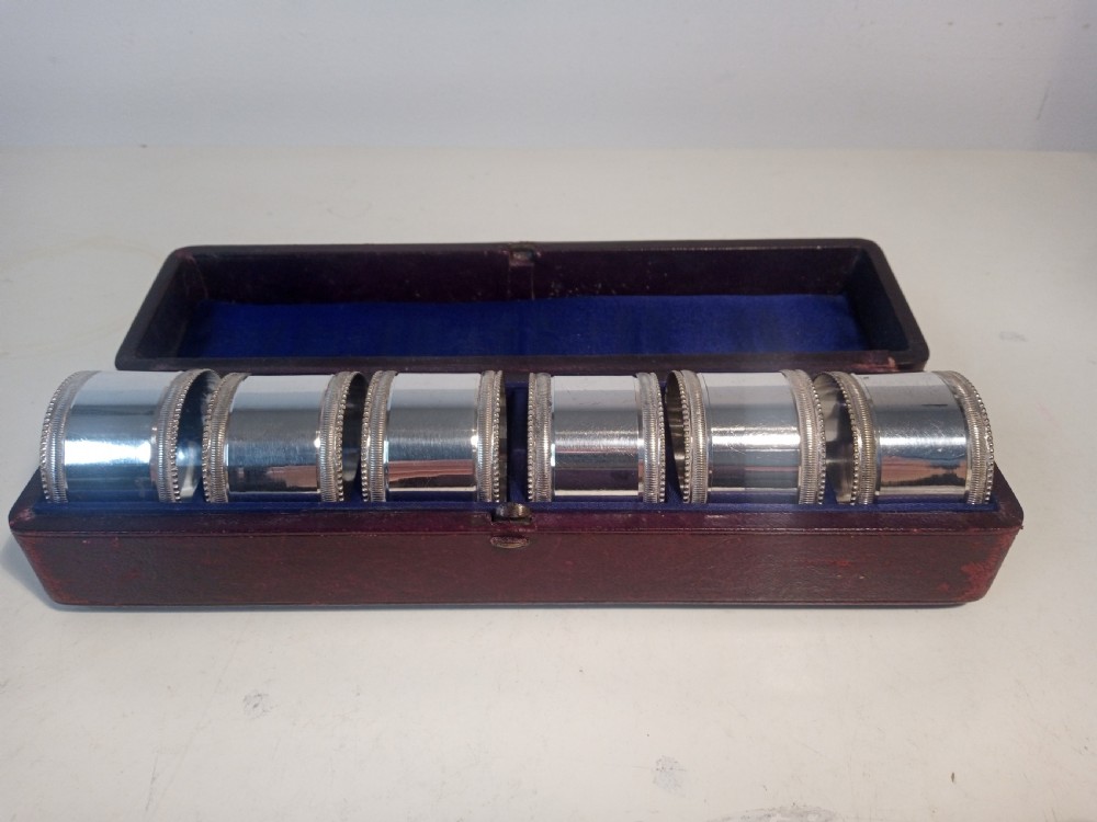 A set of six silver-plated napkin rings with beaded rims displayed inside a dark red fitted presentation case lined with blue velvet.