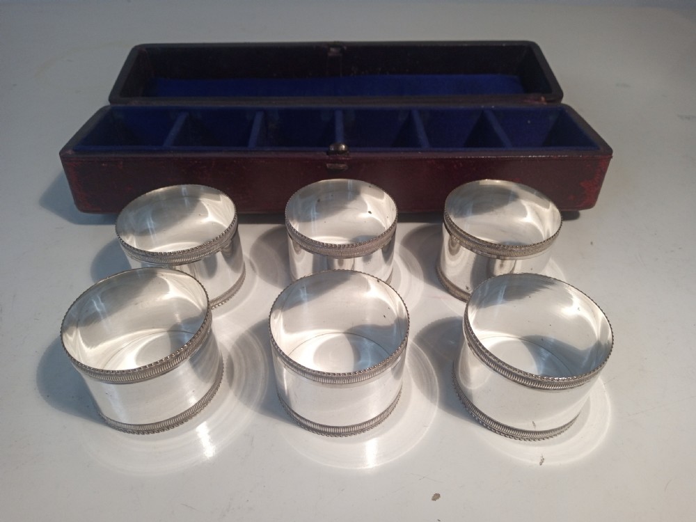 A set of six silver-plated napkin rings with beaded rims displayed inside a dark red fitted presentation case lined with blue velvet. View III