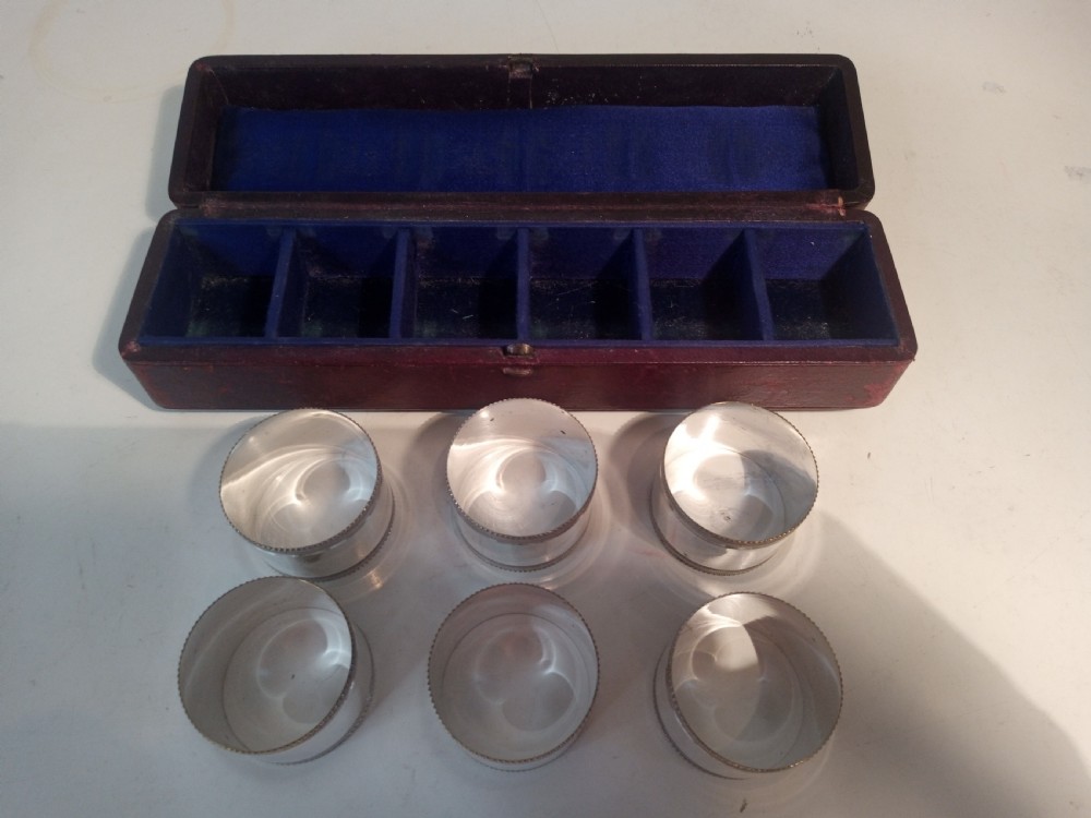 A set of six silver-plated napkin rings with beaded rims displayed inside a dark red fitted presentation case lined with blue velvet. View IV