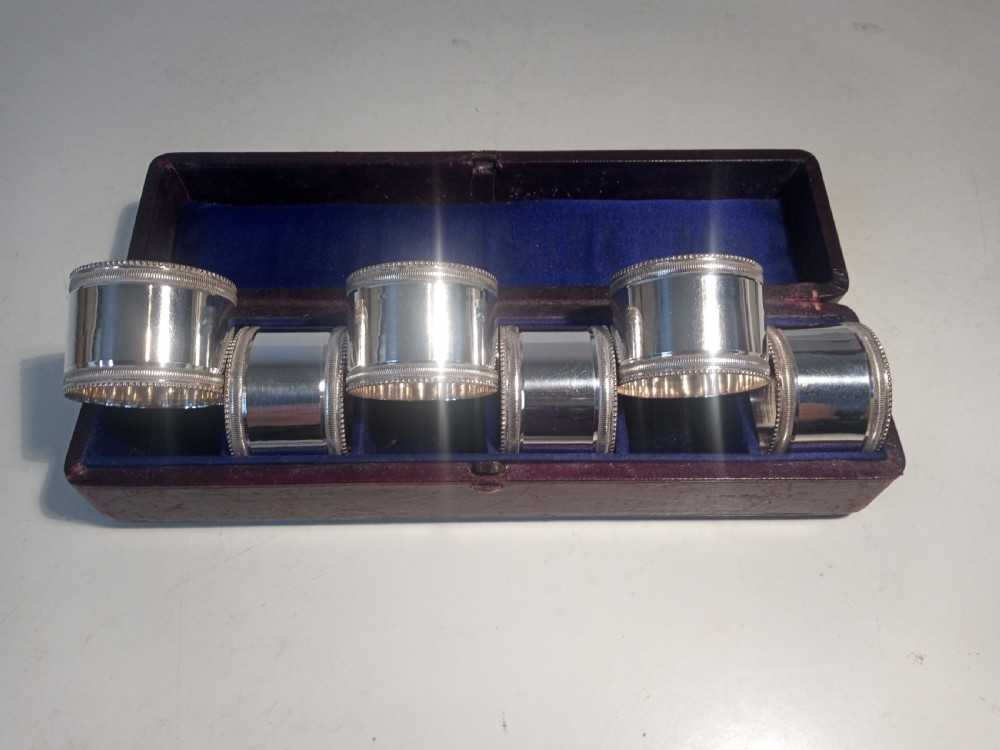 A set of six silver-plated napkin rings with beaded rims displayed inside a dark red fitted presentation case lined with blue velvet. View V