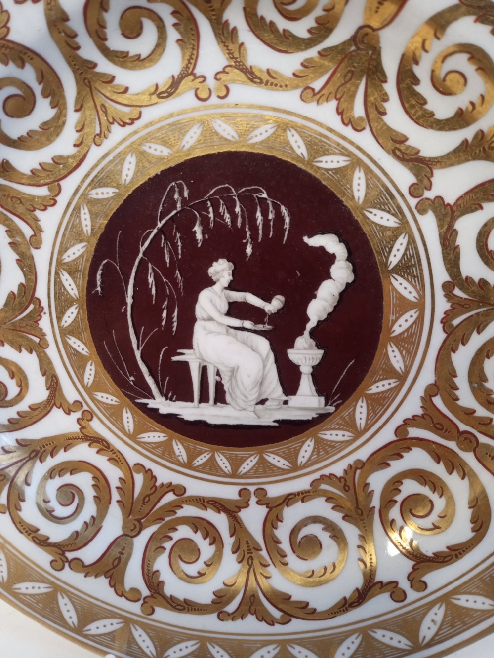 A circular Regency period porcelain plate decorated with elaborate gold scrollwork and a central maroon medallion depicting a classical female figure. View III