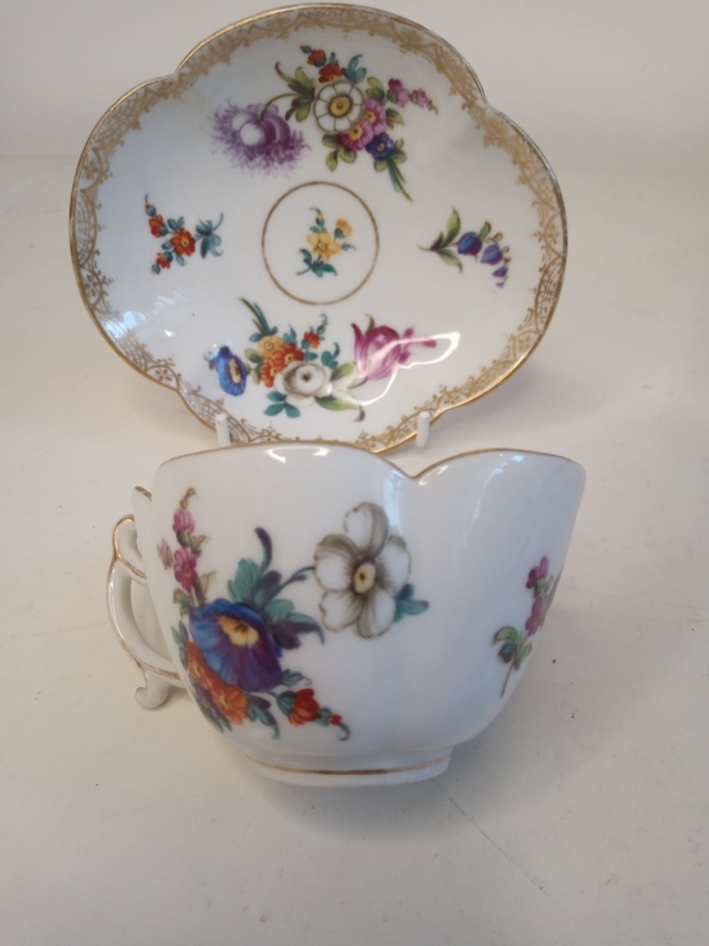 A white porcelain teacup and scalloped saucer decorated with hand-painted multicoloured floral sprays and gold gilded rims. View II