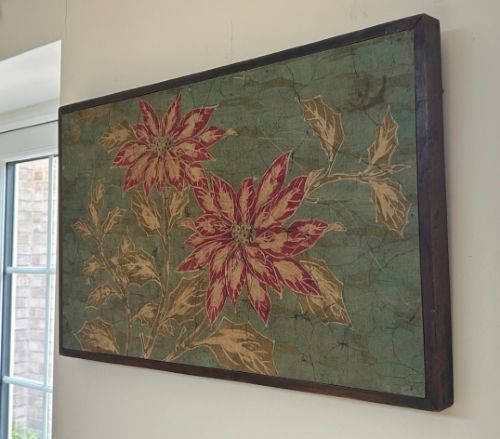 fig 1. A horizontal framed batik textile artwork displaying red and cream poinsettia flowers with foliage against a crackled sage green background.