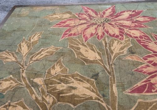 A horizontal framed batik textile artwork displaying red and cream poinsettia flowers with foliage against a crackled sage green background. View IX