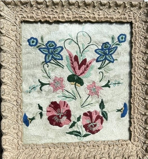 An early 20th-century framed floral embroidery panel featuring colourful hand-stitched blossoms on a cream background set within an ornate relief frame.