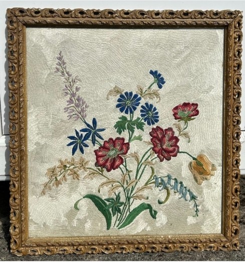A square framed needlework panel displaying a central floral arrangement in red, blue, and yellow on a cream stitched background inside a carved gold frame.