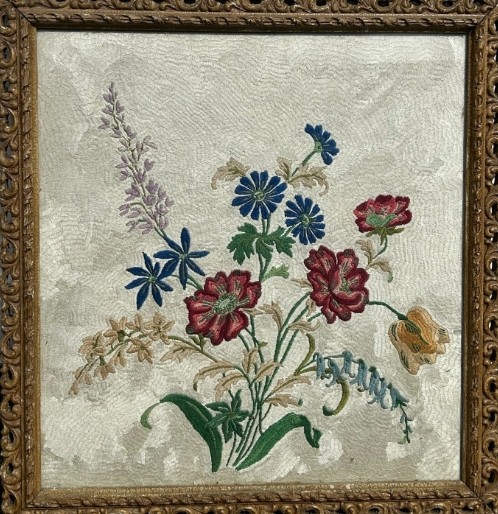 A square framed needlework panel displaying a central floral arrangement in red, blue, and yellow on a cream stitched background inside a carved gold frame. View II