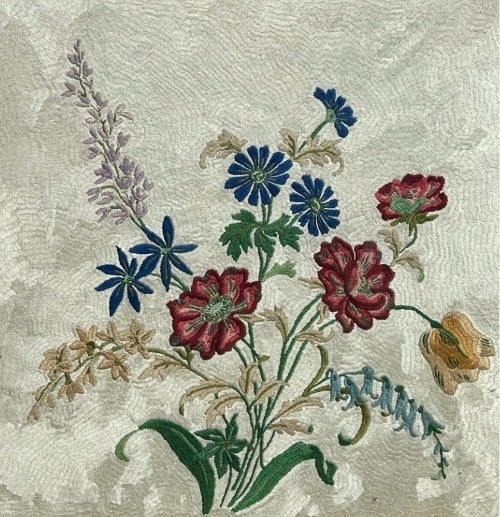 A square framed needlework panel displaying a central floral arrangement in red, blue, and yellow on a cream stitched background inside a carved gold frame. View III
