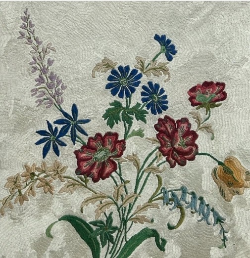 A square framed needlework panel displaying a central floral arrangement in red, blue, and yellow on a cream stitched background inside a carved gold frame. View IV