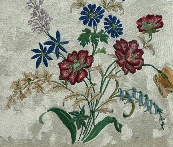 A square framed needlework panel displaying a central floral arrangement in red, blue, and yellow on a cream stitched background inside a carved gold frame. View V