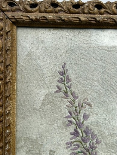 A square framed needlework panel displaying a central floral arrangement in red, blue, and yellow on a cream stitched background inside a carved gold frame. View VI