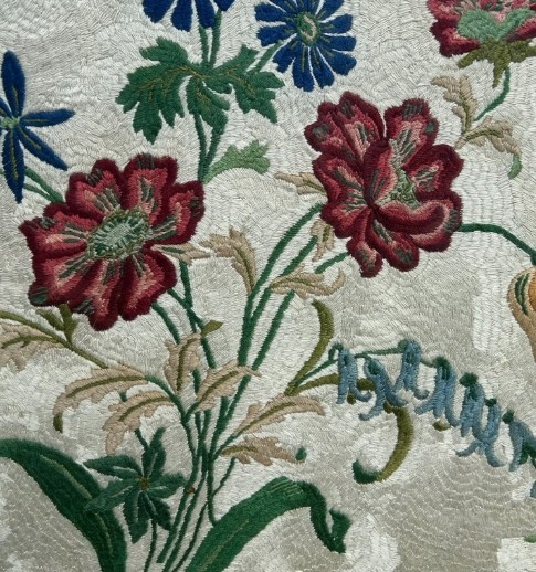 A square framed needlework panel displaying a central floral arrangement in red, blue, and yellow on a cream stitched background inside a carved gold frame. View VII