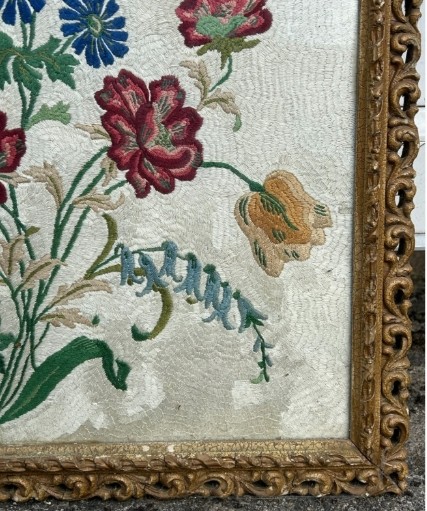 A square framed needlework panel displaying a central floral arrangement in red, blue, and yellow on a cream stitched background inside a carved gold frame. View VIII