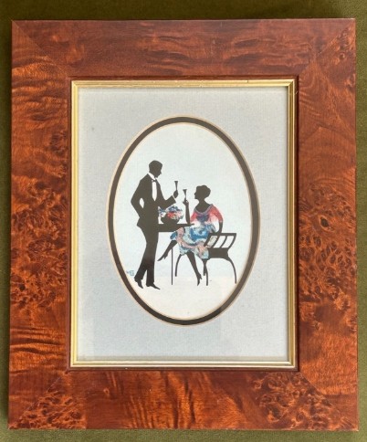 An Art Deco framed silhouette picture featuring a couple holding glasses, set within an oval mount and a wide burr wood frame.