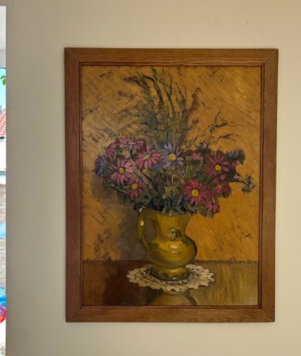 A mid-20th century oil painting on board featuring pink and purple flowers in a yellow pitcher against a textured ochre background, presented in a wooden frame. View V