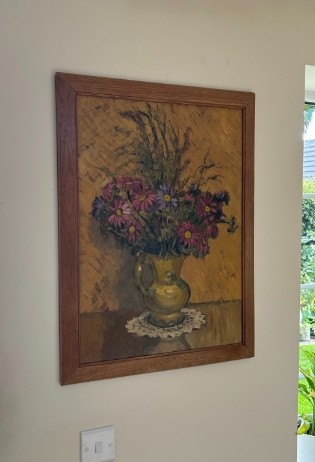 A mid-20th century oil painting on board featuring pink and purple flowers in a yellow pitcher against a textured ochre background, presented in a wooden frame. View IX