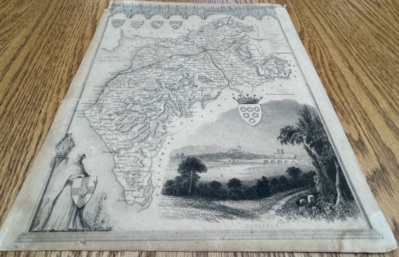 A nineteenth-century monochrome steel engraving map of Cumberland by Thomas Moule featuring decorative Gothic tracery borders, coats of arms, and an architectural vignette of Carlisle. View II