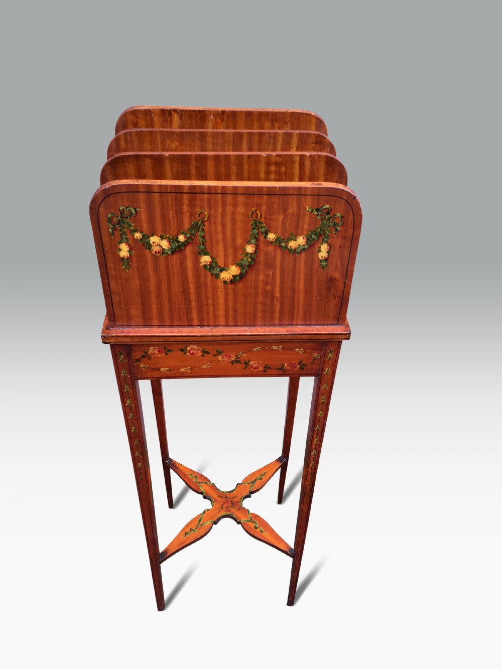 Late 19th-century satinwood standing bookstand with hand-painted rose garlands, vertical book divisions, tapered legs, and an X-shaped stretcher.