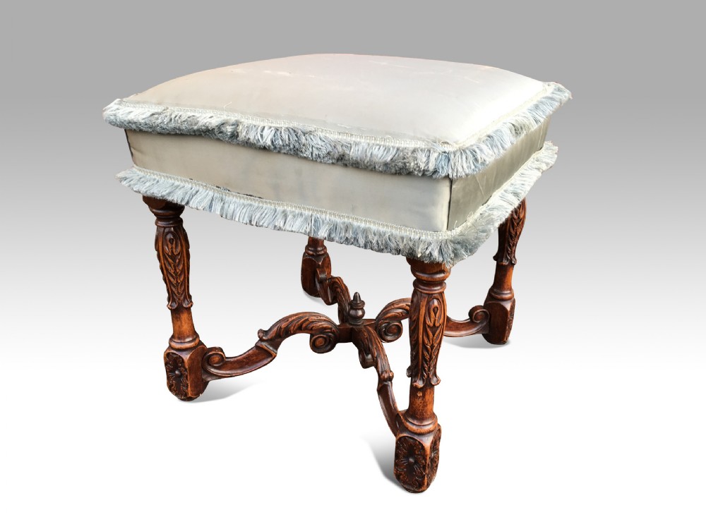 A carved walnut stool featuring a pale blue upholstered cushion with fringe trimming, supported by carved legs and an X-frame stretcher.