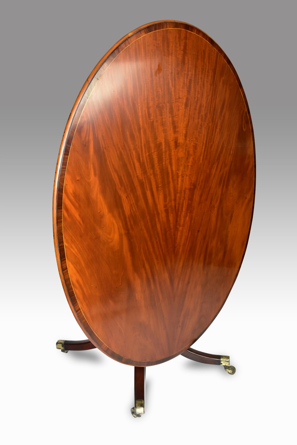 A George III oval mahogany tilt-top breakfast table with crossbanded top and splayed legs on brass castors, shown tilted vertically.