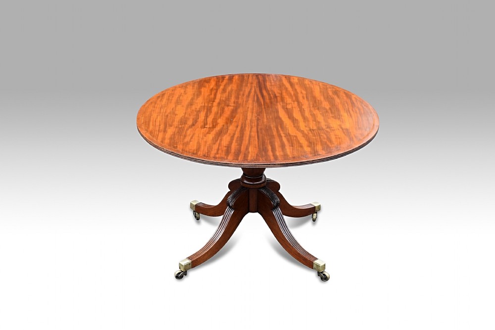 A circular Regency mahogany centre table with lacewood banding, resting on a central pedestal with four carved reeded legs and brass castors.