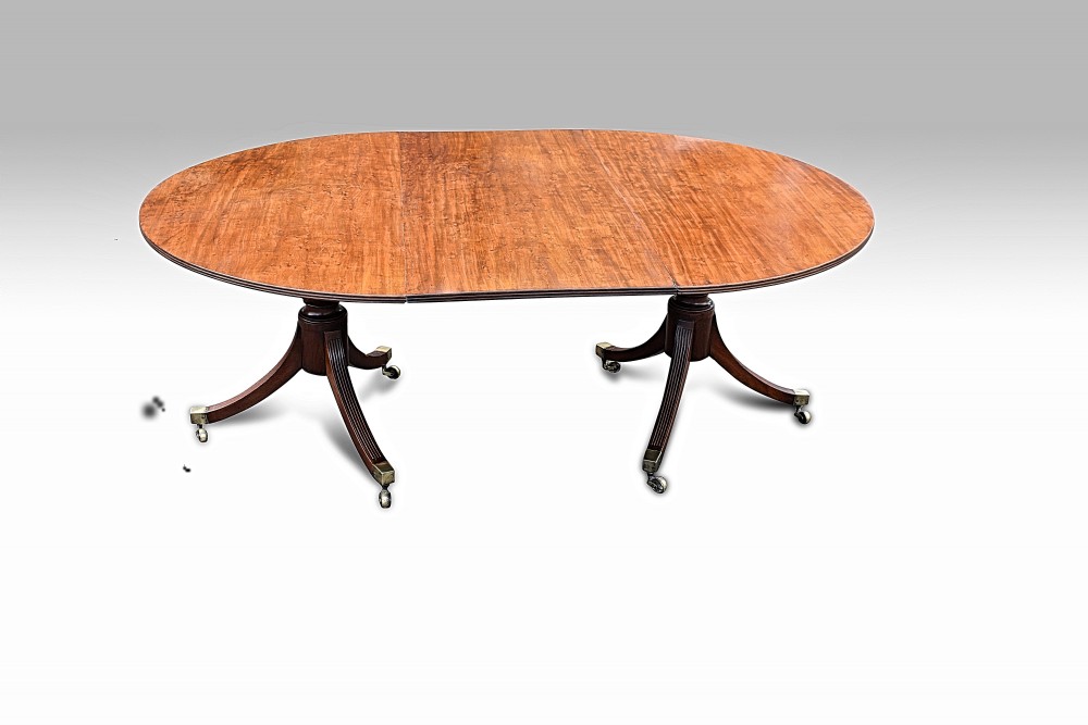 An antique George III oval mahogany dining table featuring a twin pillar pedestal base, reeded legs, and brass castors.